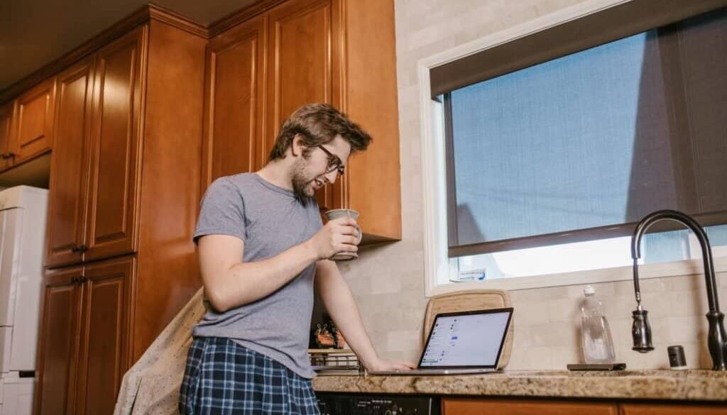 Man in pajamas enjoying coffee while browsing a laptop in a kitchen setting.