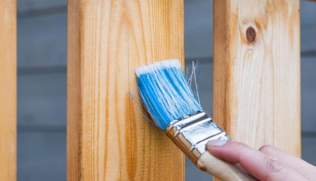 A hand painting a wooden fence plank with a blue paintbrush outdoors. Perfect for DIY and renovation themes.