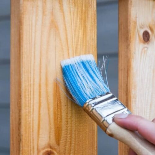 A hand painting a wooden fence plank with a blue paintbrush outdoors. Perfect for DIY and renovation themes.
