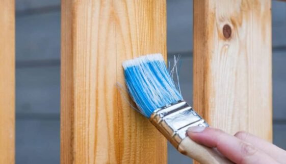 A hand painting a wooden fence plank with a blue paintbrush outdoors. Perfect for DIY and renovation themes.